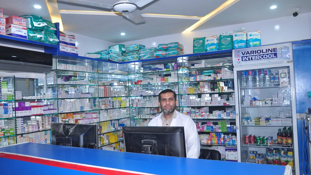 A pharmacist behind the counter of the Asaaf Hospital pharmacy in Pakistan, with shelves of medicine packs that will now carry mandatory 2D barcodes