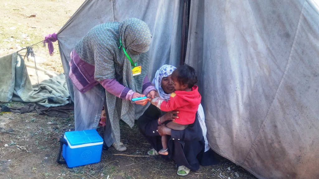 A health worker marking a young child's finger after polio vaccination outside a tent in Pakistan, with a blue vaccine cold box beside her