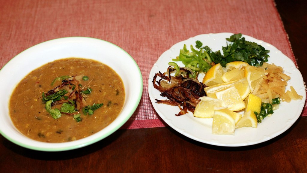A bowl of Pakistani haleem topped with fried onions and coriander, served with lemon wedges, ginger, green chillies and herbs, the garnishes in this haleem recipe