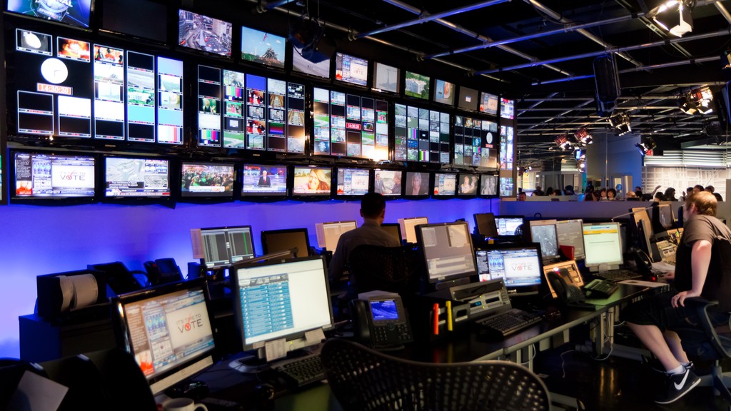 A television broadcast control room with a wall of live feed monitors above newsroom desks and operators