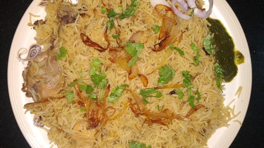 A plate of chicken yakhni pulao with fluffy basmati rice, a chicken piece, fried onions, fresh coriander, onion rings and green chutney