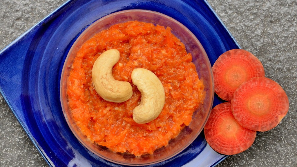 A bowl of gajar ka halwa topped with cashew nuts, with fresh red carrot slices beside it on a blue plate
